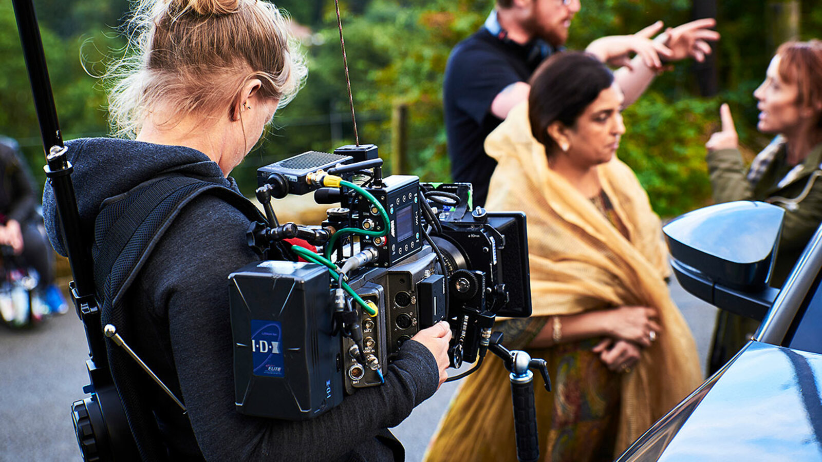 A camera operator is pictured from behind, holding a camera towards two people that are being directed for a shot.