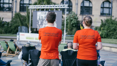 ShAFF volunteers Anna and Phil stand with the ir backs to the camera watching an outdoor screen in Sheffield's Peace Gardens