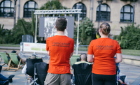 ShAFF volunteers Anna and Phil stand with the ir backs to the camera watching an outdoor screen in Sheffield's Peace Gardens