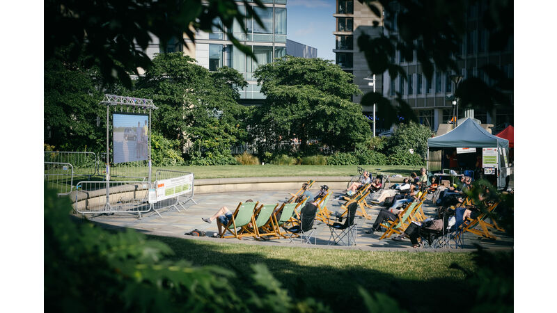 An outdoor cinema screen in Sheffield's Peace Gardens, rows of audience members are seated in deckchairs to watch the films