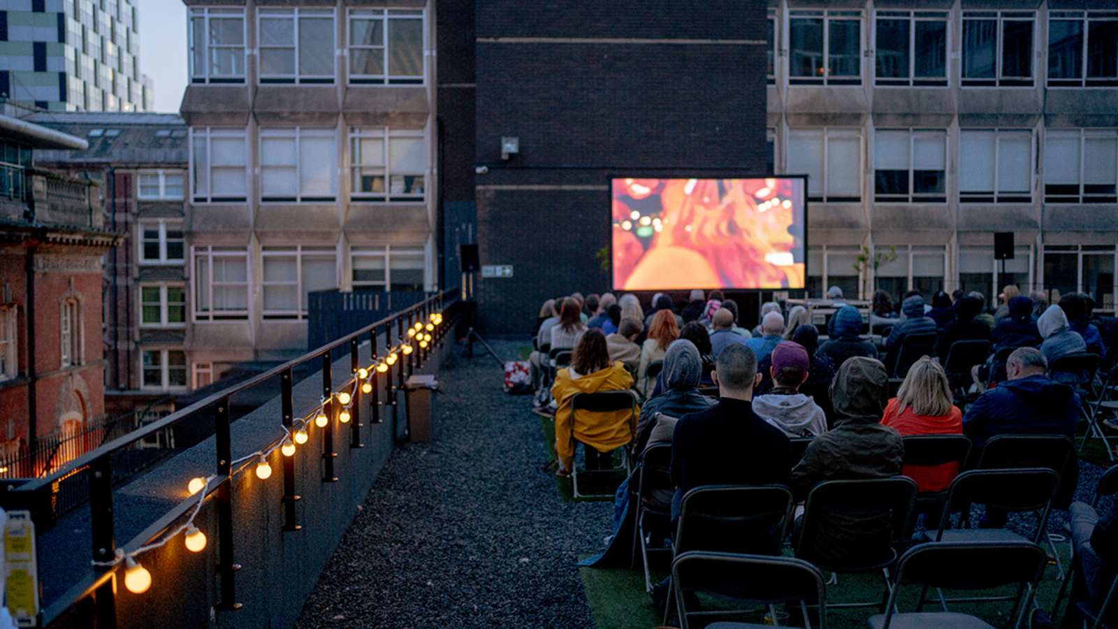 A group of people sat on a rooftop watching a film on a large screen