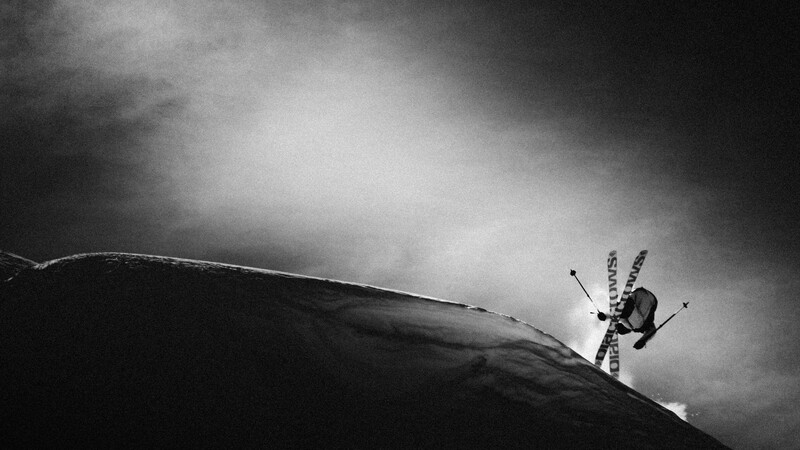 black and white image of a skier crossing skis in a trick, lit by a shaft of light in the darkness