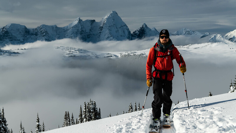 A skier skiing towards the camera above a coud inversion with jagged snowy peaks in the background.