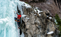 An climber wearing red climbing a dramatic frozen waterfall using ice axes.