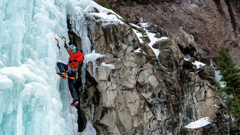 An climber wearing red climbing a dramatic frozen waterfall using ice axes.