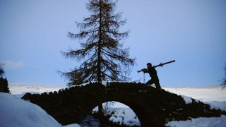 A dark figure is outlined as they carry skis across a small bridge throguh a snowy landscape past a lone tree