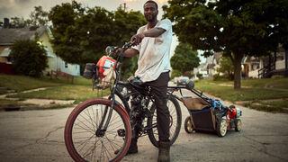 A Black man stands astride a bicycle which has a lawnmower attached to the back of it