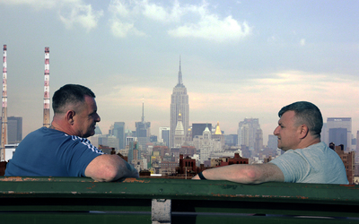 two men sit facing each other on a bench, with a cityscape in the distance between them
