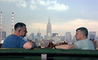 two men sit facing each other on a bench, with a cityscape in the distance between them