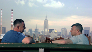 two men sit facing each other on a bench, with a cityscape in the distance between them
