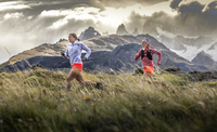 Two women run at speed across a grassy landscape, snowy moutains in the distance behind them