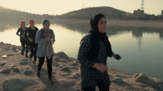 A group of four Afghan women run on rocky ground beside a lake, they are wearing headscarves, trail shoes and running packs.