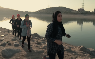 A group of four Afghan women run on rocky ground beside a lake, they are wearing headscarves, trail shoes and running packs.
