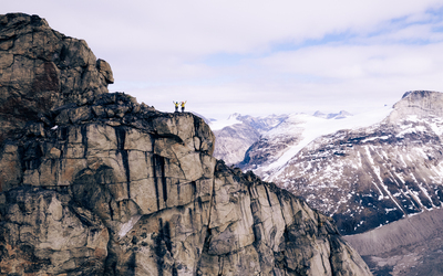 Two adventurers stand side by side in the distance raising their arms to the camera, they are dwarfed by the rocky landscape that surrounds them.