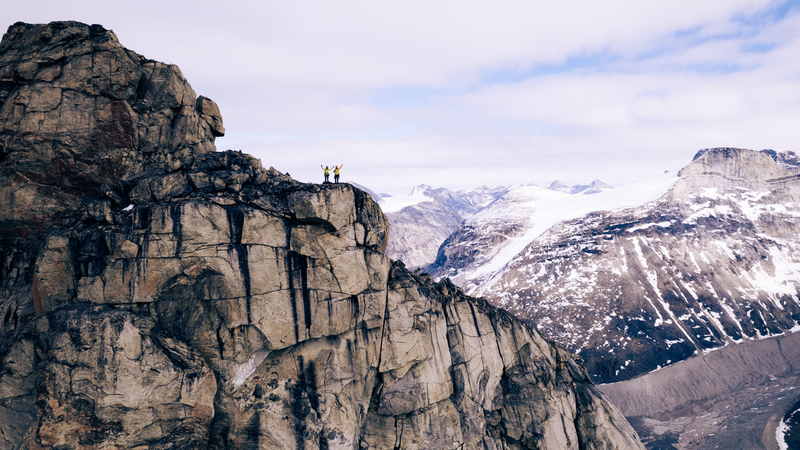 Two adventurers stand side by side in the distance raising their arms to the camera, they are dwarfed by the rocky landscape that surrounds them.