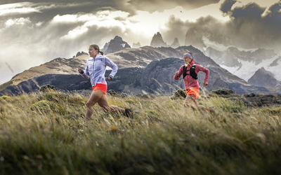 Two women run at speed across a grassy landscape, snowy moutains in the distance behind them