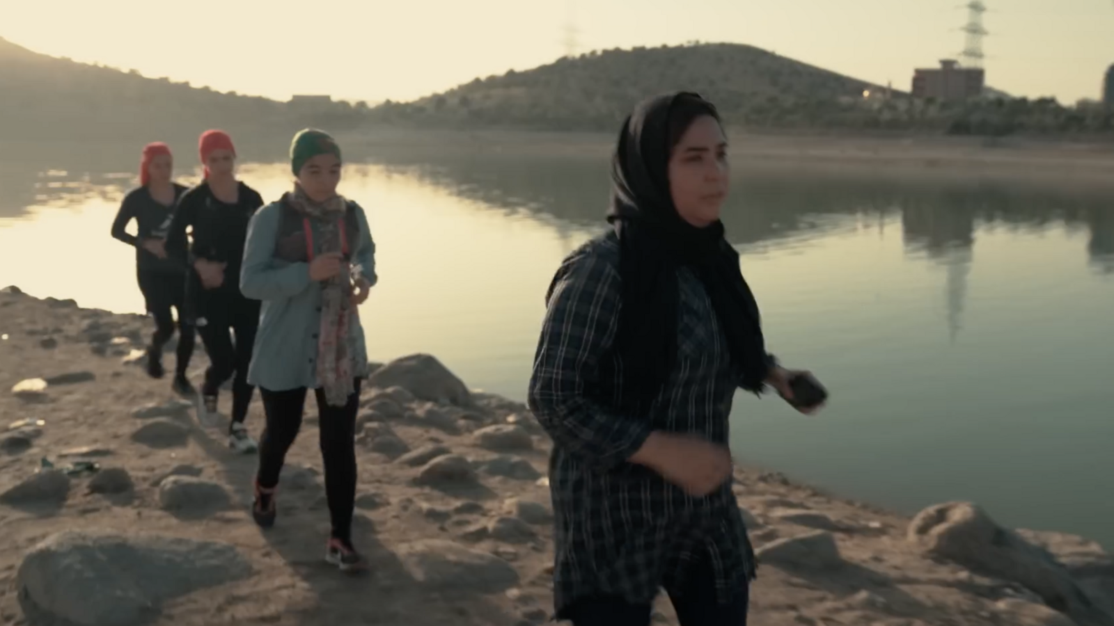 A group of four Afghan women run on rocky ground beside a lake, they are wearing headscarves, trail shoes and running packs.