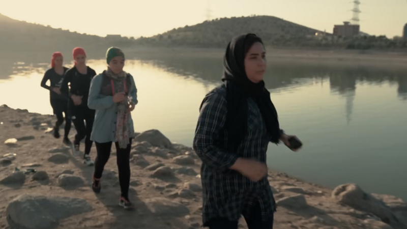 A group of four Afghan women run on rocky ground beside a lake, they are wearing headscarves, trail shoes and running packs.