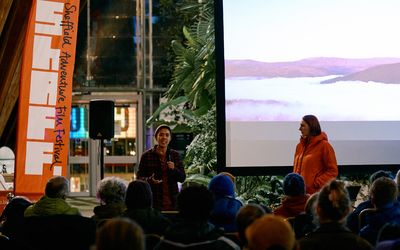 Speaker Frit Tam talks to an audience while standing in front of a cinema screen, ShAFF codirector Anna Paxton stands next to him listening
