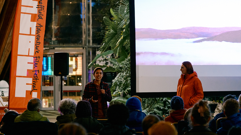 Speaker Frit Tam talks to an audience while standing in front of a cinema screen, ShAFF codirector Anna Paxton stands next to him listening