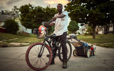 A Black man stands astride a bicycle which has a lawnmower attached to the back of it