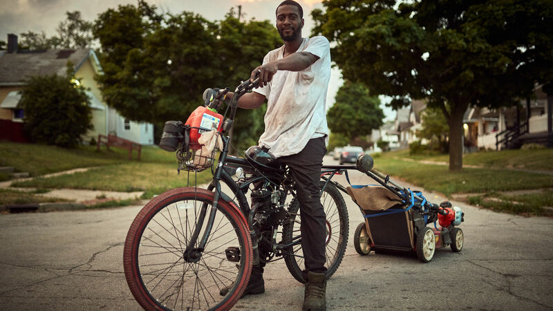 A Black man stands astride a bicycle which has a lawnmower attached to the back of it