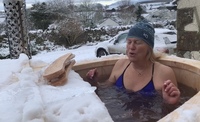 A woman sits in a cold water tub surrounded by snow, she is closing her eyes and exhaling with the cold