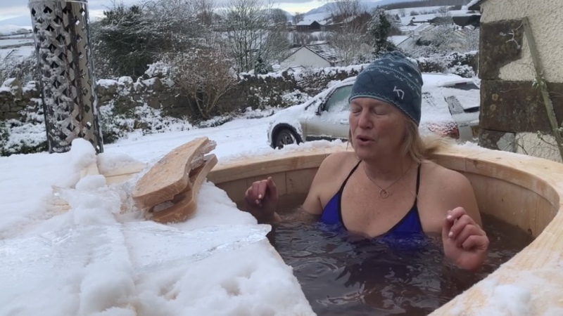 A woman sits in a cold water tub surrounded by snow, she is closing her eyes and exhaling with the cold