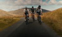 Three cyclists ride side by side along a narrow strip of road, surrounded by grassy Welsh hills