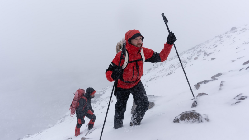 A white man, wearing black snow trousers and a red snow jacket with black segments on the elbows, makes his way uphill through rocky terrain covered in snow, using walking poles. He looks forwards, at the way ahead. He is wearing a white rucksack with a rope attached to the top. Behind him, another figure follows him uphill, also carrying a rucksack with a rope on top. The weather looks windy and cold, with snow swirling around them. Beyond the hillside, everything is grey, the view blocked by the weather.