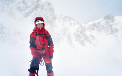 A white woman, wearing matching red snow trousers and jacket, with black segments on the elbows and knees, and a harness, stands in the snow, holding an ice axe with one hand and a snow white helmet on top of her head. The hood of her snow jacket is up, and her firey hair pokes out, caught in the wind. Snow swirls up around her, and behind her looms a bright snowy mountain face. The sky is light grey and snowy.