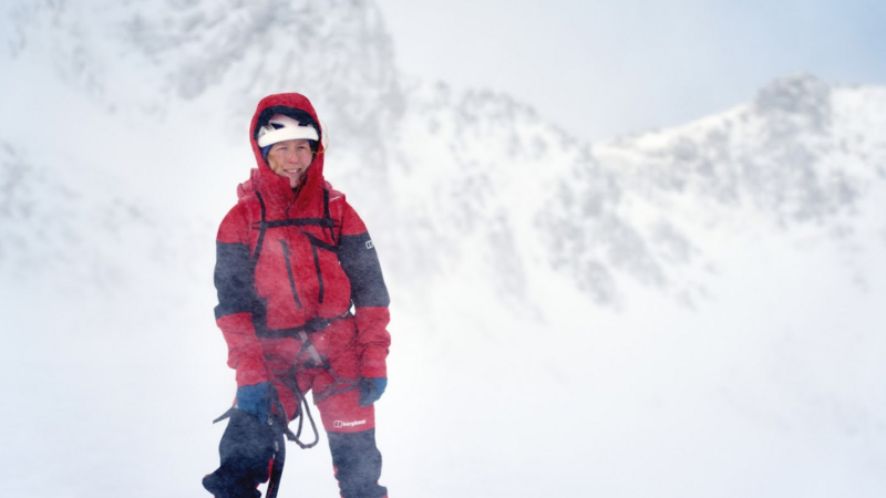 A white woman, wearing matching red snow trousers and jacket, with black segments on the elbows and knees, and a harness, stands in the snow, holding an ice axe with one hand and a snow white helmet on top of her head. The hood of her snow jacket is up, and her firey hair pokes out, caught in the wind. Snow swirls up around her, and behind her looms a bright snowy mountain face. The sky is light grey and snowy.