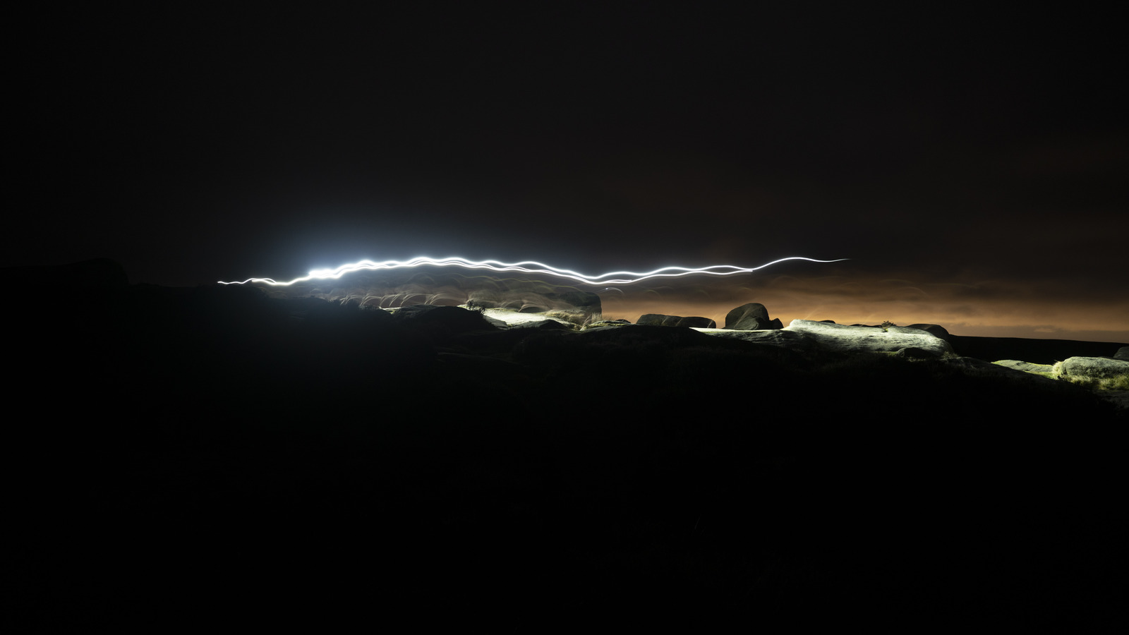 In the dark, a headtorch trail bounces across the frame, lighting up part of a rocky peak clifftop, and creating a white streak in the dark.