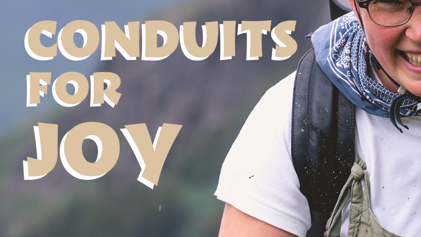 A front-on mid-close up of a white non-binary person called Ali, wearing a bright white tee, khaki dungarees, a multicoloured cycling cap, and a dark helmet. They have a navy bandana tied around their neck, dark rimmed glasses, and a huge grin across their face. The black strap of their bassoon rucksack case is spattered with mud. Only half of them can be seen - the other half off screen. Behind them is a green blur of overlapping hillsides. On top of this backdrop are the words "Conduits for Joy" in beige, with a white block shadow. 