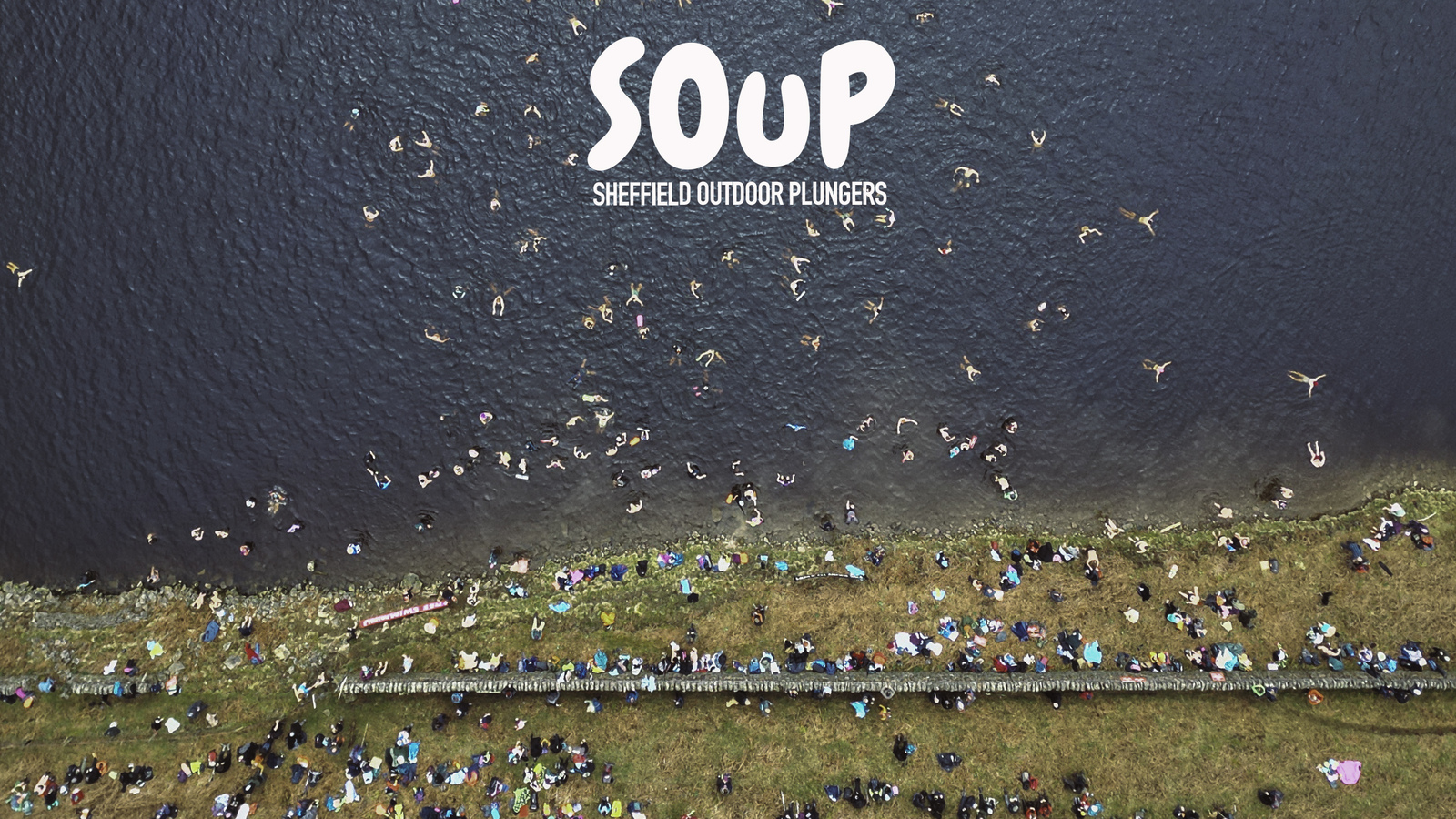 Bird's eye aerial view of numerous people swimming in a Peak District reservoir, with many more standing at the water's edge.