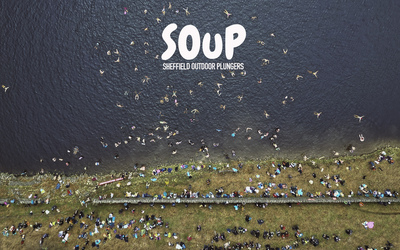 Bird's eye aerial view of numerous people swimming in a Peak District reservoir, with many more standing at the water's edge.