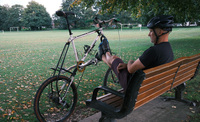 Jon rests his feet on his tall bike whilst sitting on a park bench