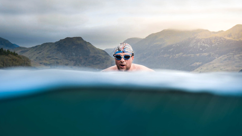 A white man swims in a lake with mountains behind