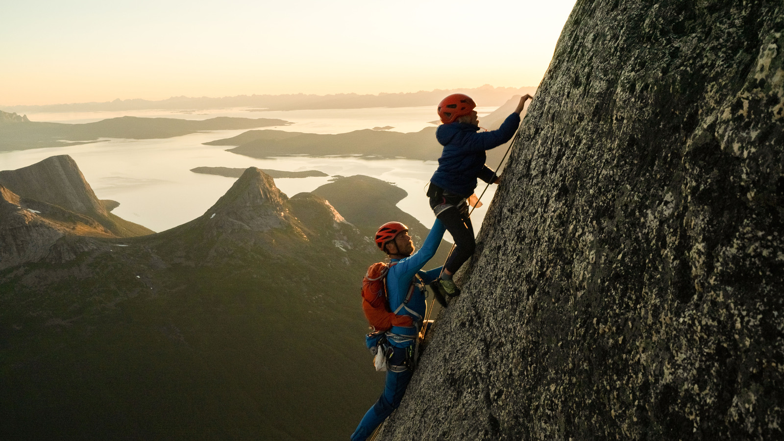 Freya, A 9 year old girl climbs a huge granite gliff ahead of her father, leo houlding. They are silhouetted by the low summer evening sun.