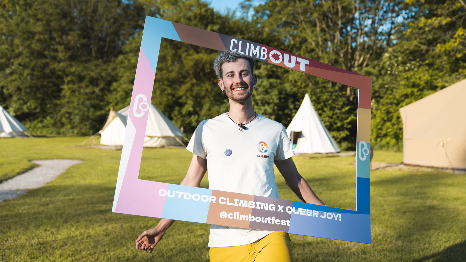 AJ is seen smiling holding a cut out rainbow-coloured frame on their head with the word "ClimbOut" on