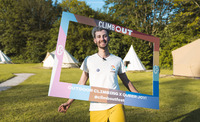 AJ is seen smiling holding a cut out rainbow-coloured frame on their head with the word "ClimbOut" on