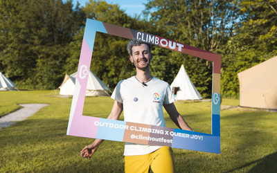 AJ is seen smiling holding a cut out rainbow-coloured frame on their head with the word "ClimbOut" on