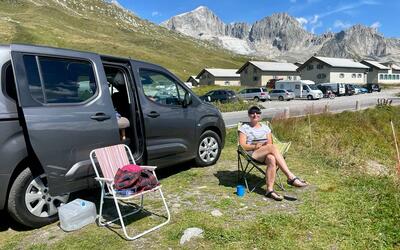 Sophie is sat beside her e-camper van in a camping chair, smiling at the camera. It's sunny and there are mountains behind her in the distance 