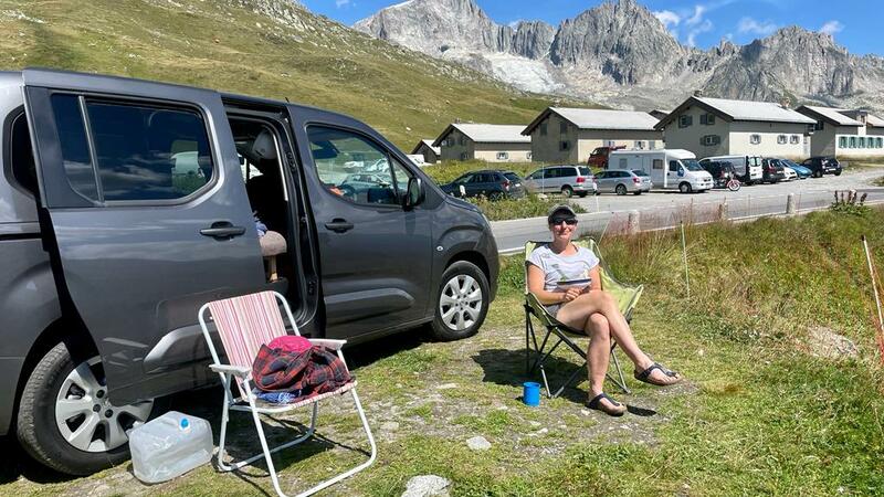 Sophie is sat beside her e-camper van in a camping chair, smiling at the camera. It's sunny and there are mountains behind her in the distance 