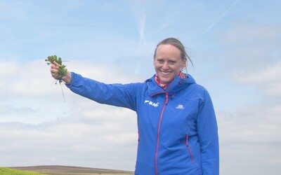 Catherine is in a blue waterproof jacket with a smile on her face, holding up moss in her hand