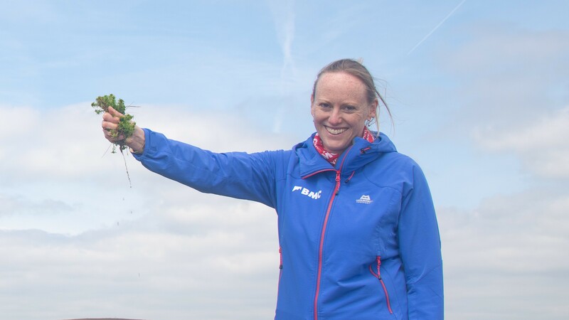 Catherine is in a blue waterproof jacket with a smile on her face, holding up moss in her hand