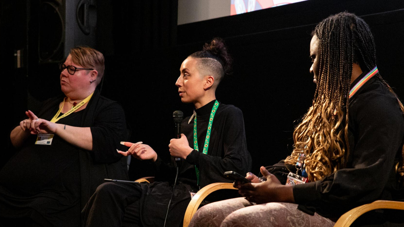 Three people sit on stage taking part in a panel discussion. One is holding a microphone and the other is doing British Sign Language.