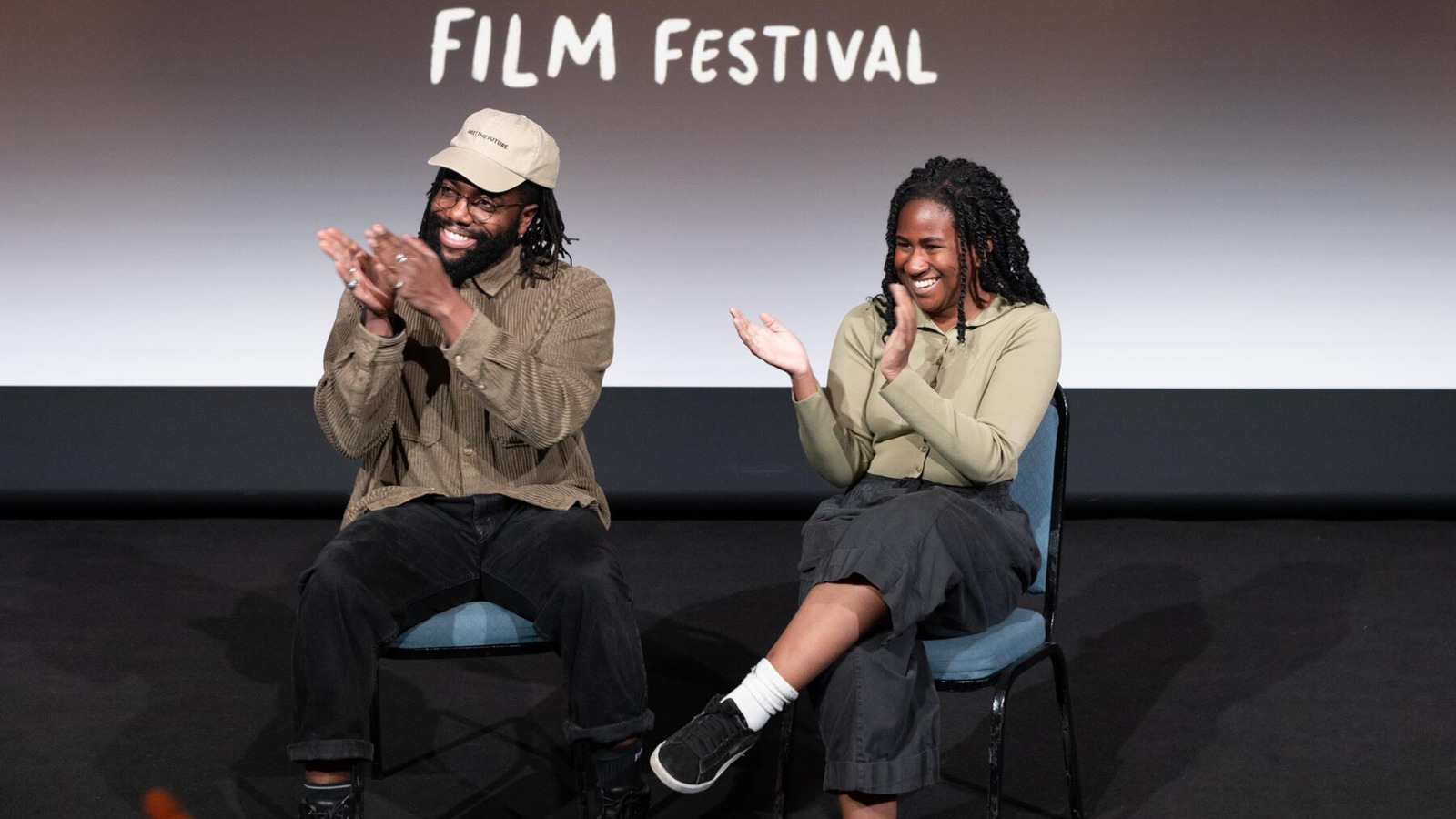 Two panel speakers are sat on stage clapping and smiling. Behind them, on the cinema screen, text reads: Film Festival.