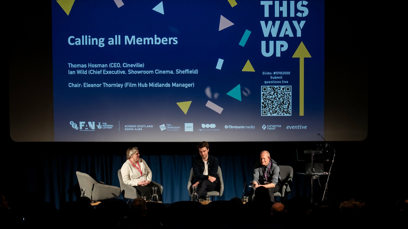 Three panel speakers sit in armchairs on a cinema theatre stage. Behind them, a slide on screen reads: This Way Up, Calling all Members