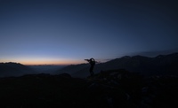 Young mountain bike athlete Bo Schwarz carries a mountain bike on his back. Wearing a bright headlamp he is silhouetted with the smooth morning colors of blue and orange filling the mountain landscape behind him.